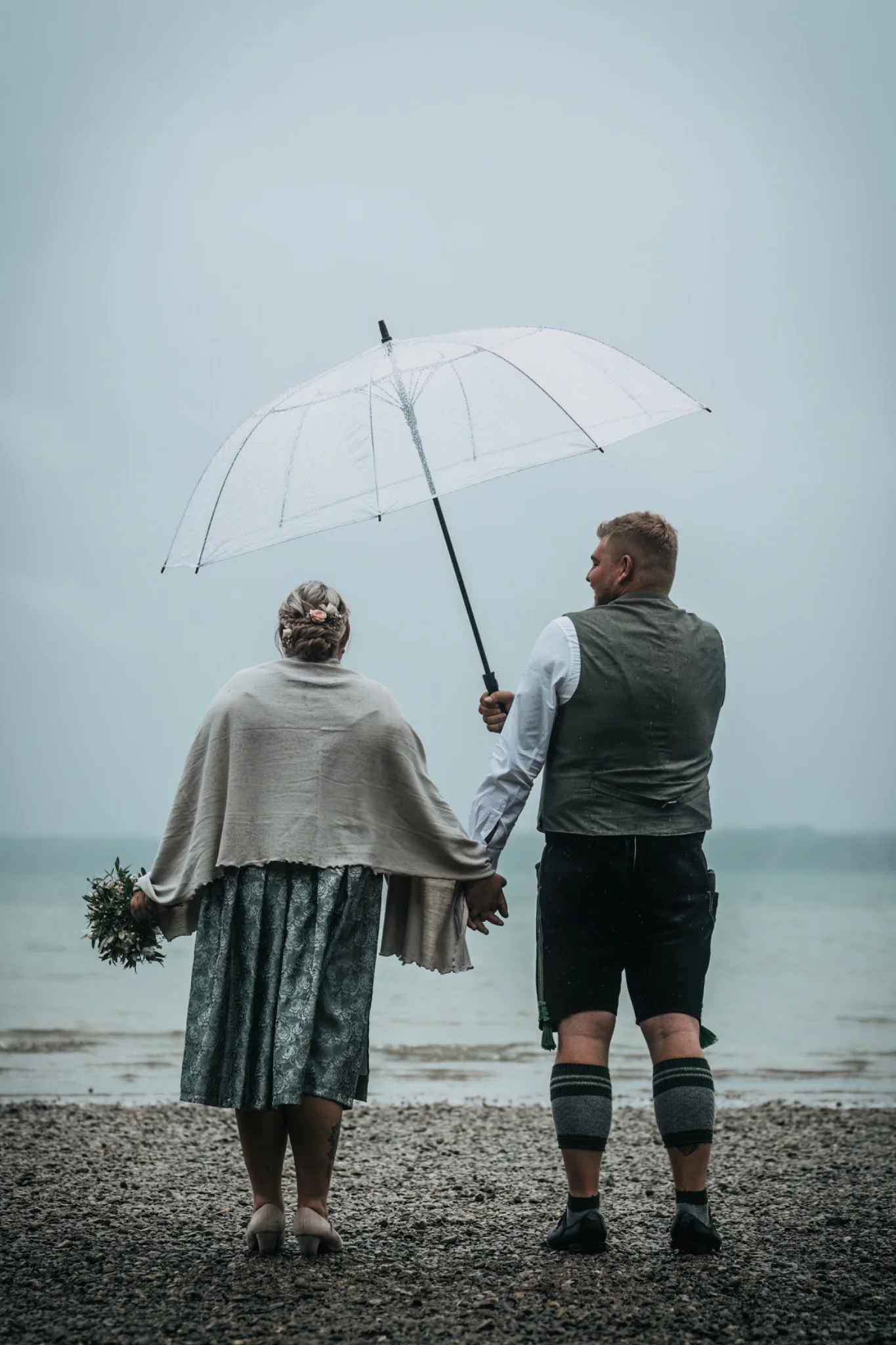 Brautpaar mit transparentem Regenschirm am See – natürliche Hochzeitsfotografie bei Regen