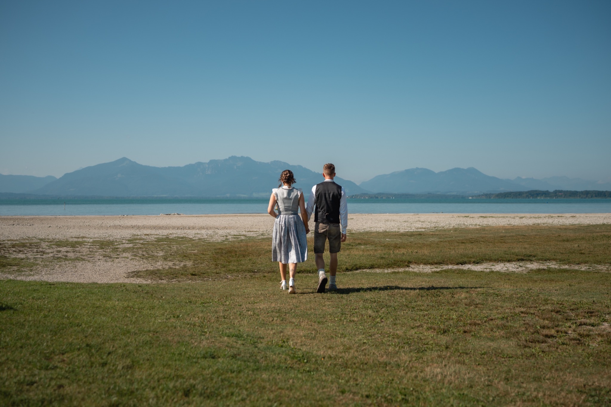 Brautpaar in Tracht spaziert Hand in Hand am See mit Bergpanorama