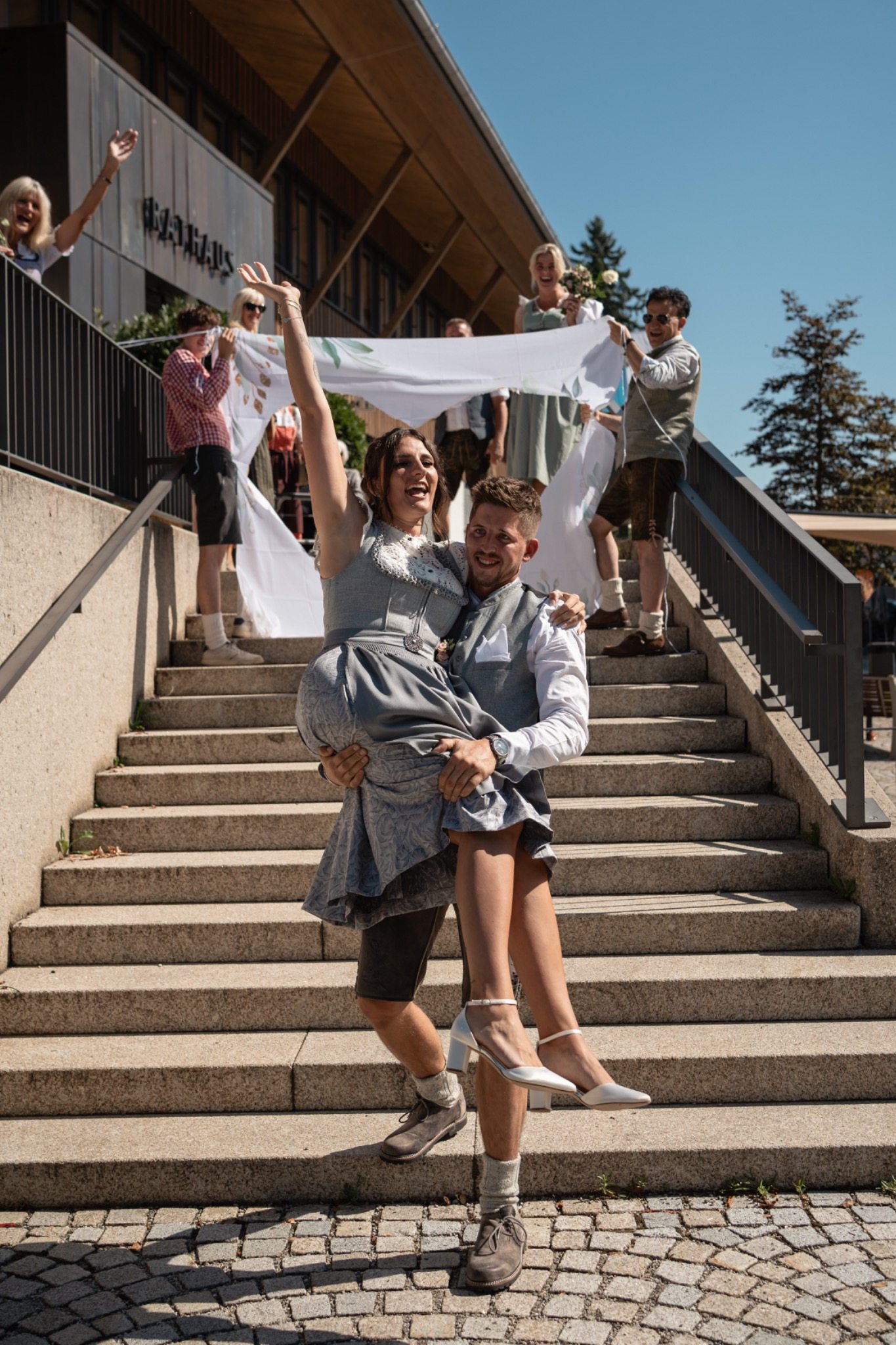 Fröhliches Brautpaar in Tracht auf einer Treppe, umgeben von jubelnden Hochzeitsgästen
