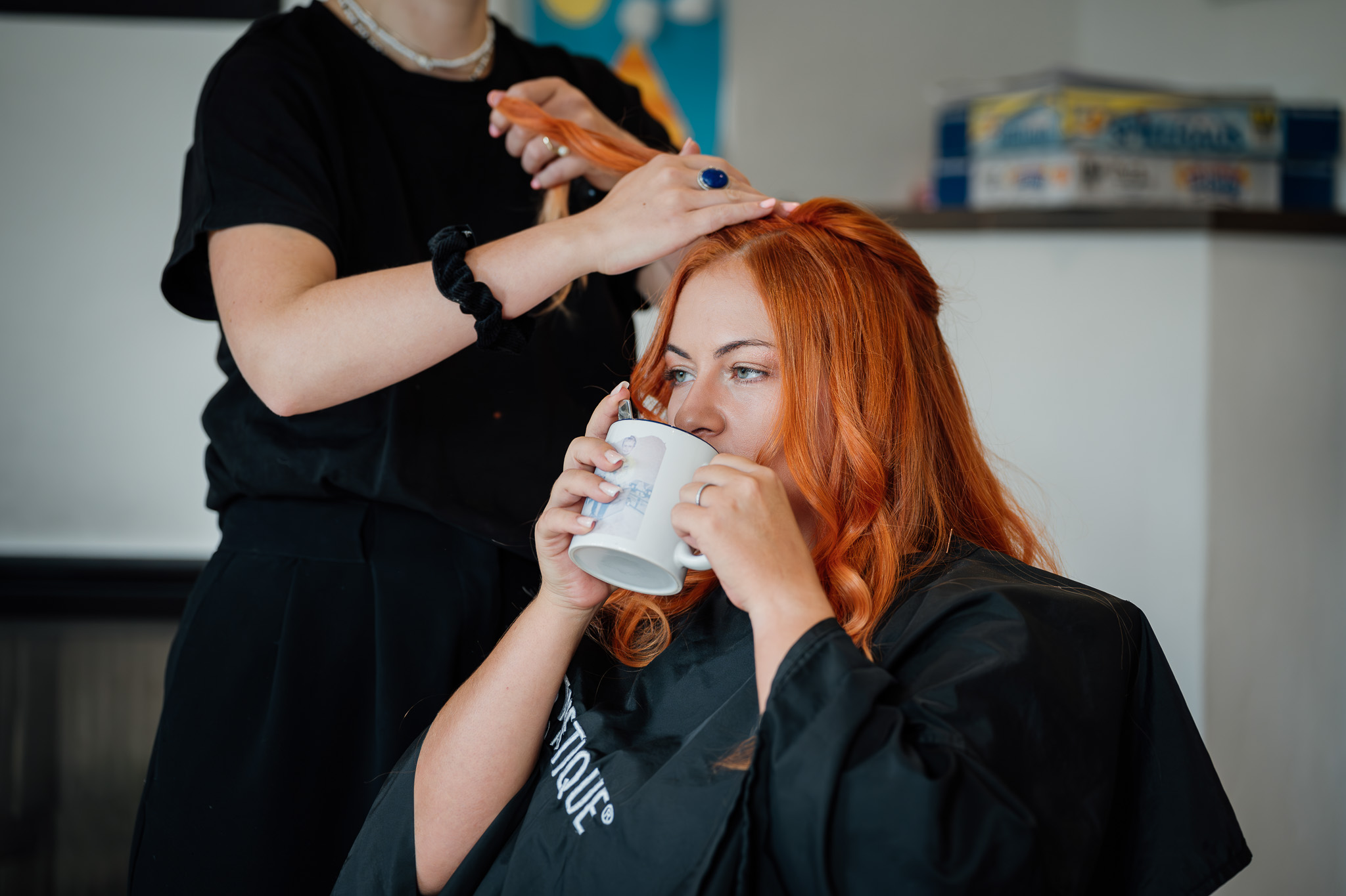 Die Braut genießt beim Getting Ready einen Kaffee, während die Stylistin an ihrer Frisur arbeitet.